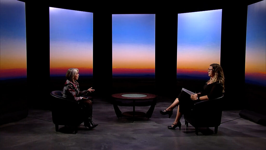 Two women sitting in chairs in front of a large screen.