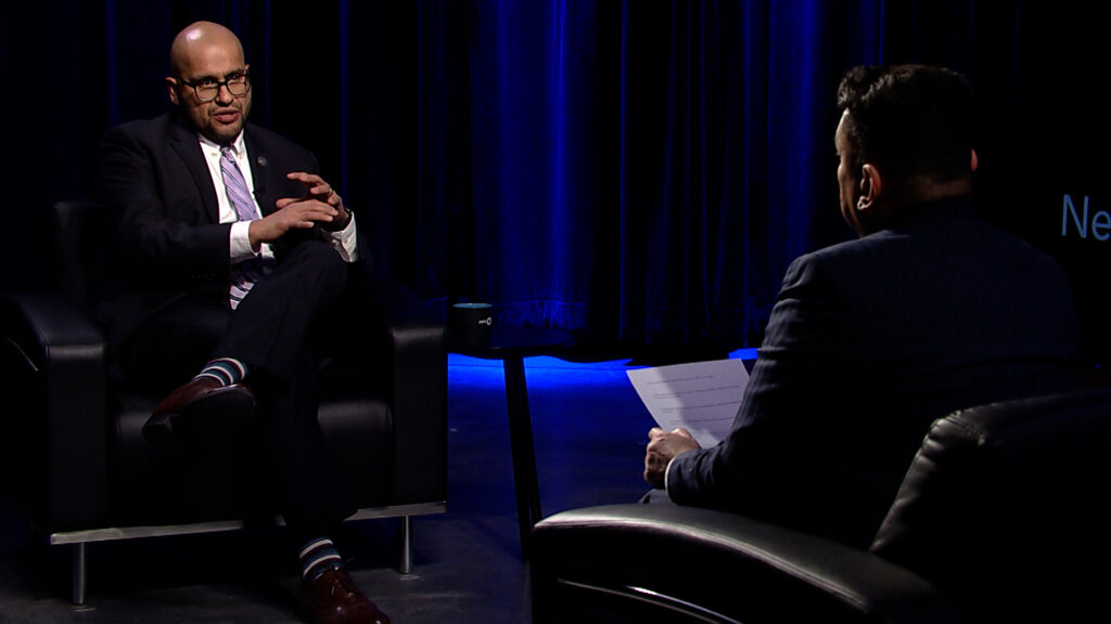 A man in a suit sitting in a chair talking to another man.