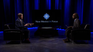 Two men sitting in chairs in front of a blue screen.
