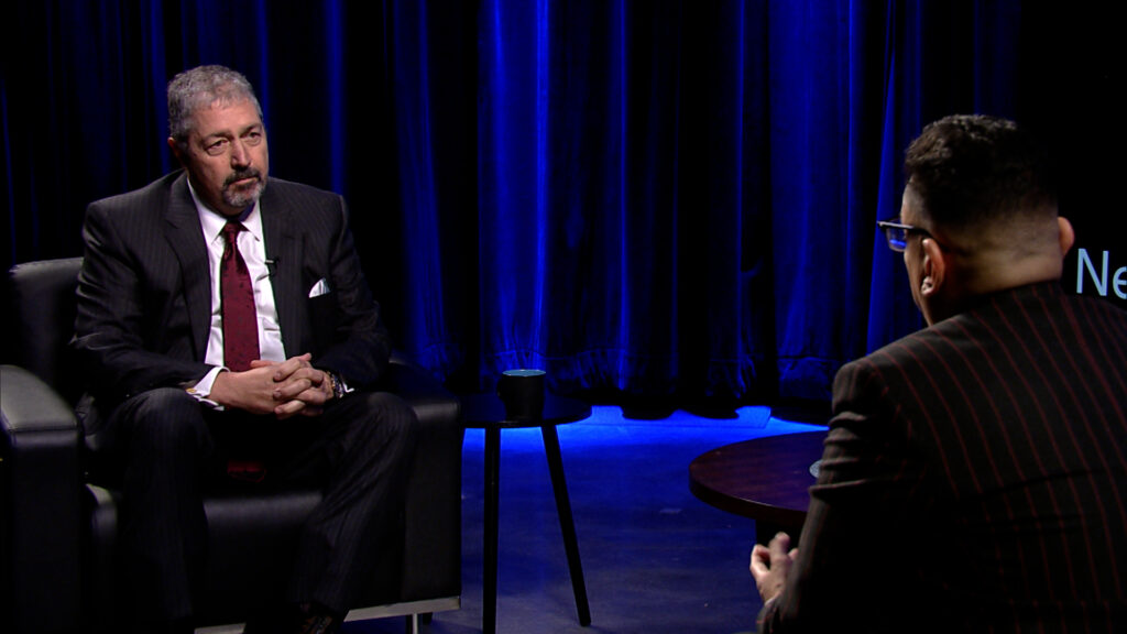 A man in a suit sits in a chair and talks to another man.