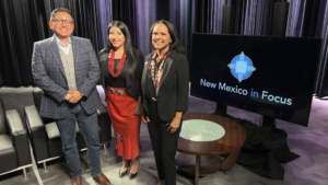 Three people standing in front of a tv with the words new mexico in focus.
