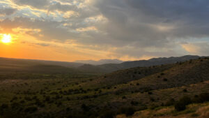 The sun is setting over a valley with mountains and water in the background.