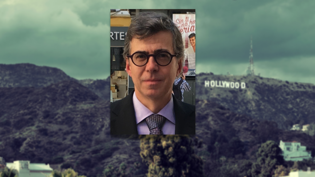 An image of a man with glasses in front of a hollywood sign.