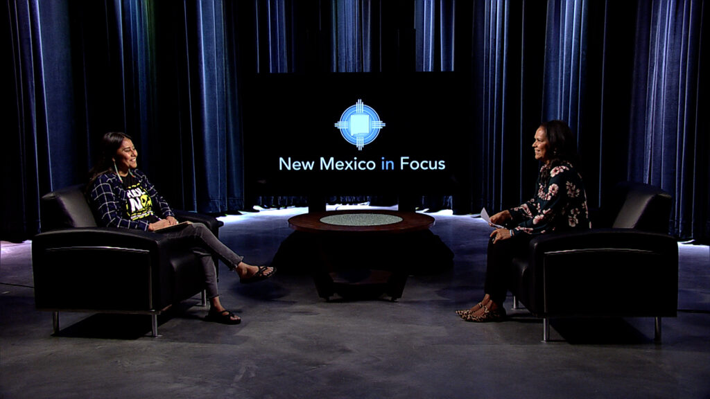 Two women sitting in chairs on a stage with the words new mexico focus.