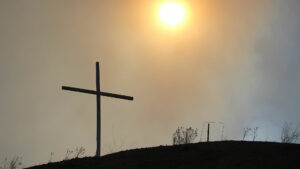 A cross sits on top of a hill with the sun behind it.