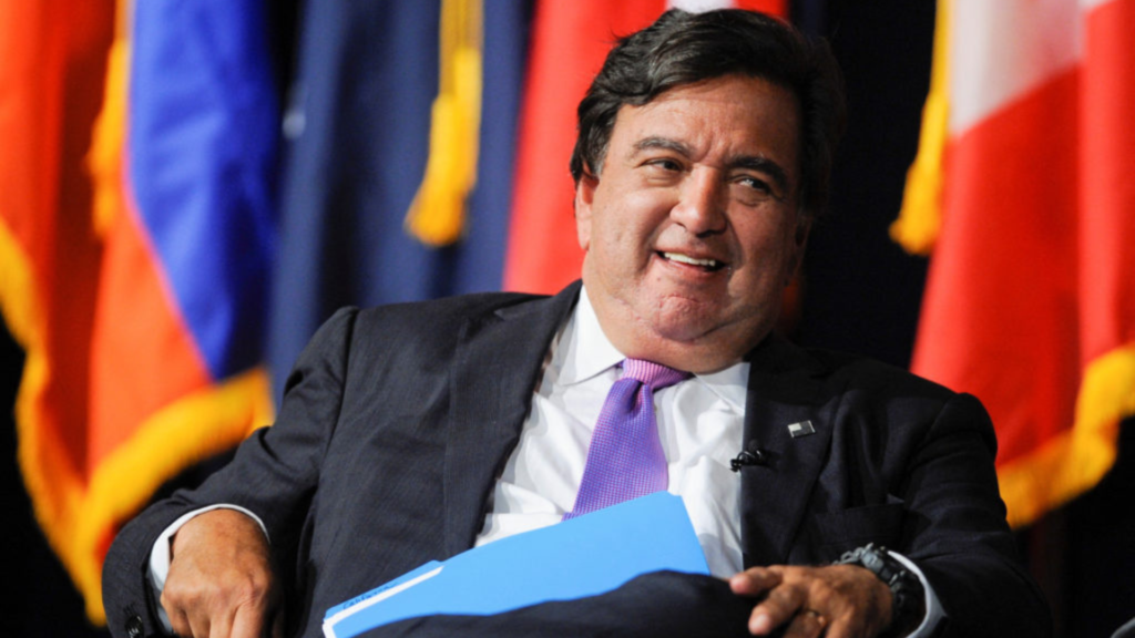 A man in a suit and tie sitting in front of flags.