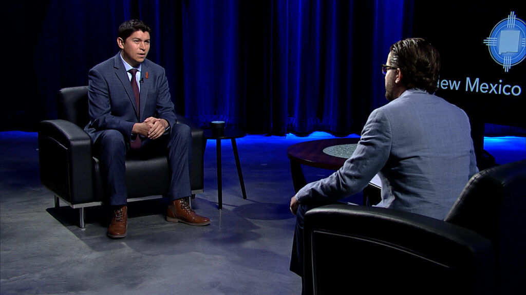 A man in a suit sits in a chair and talks to a man in a suit.