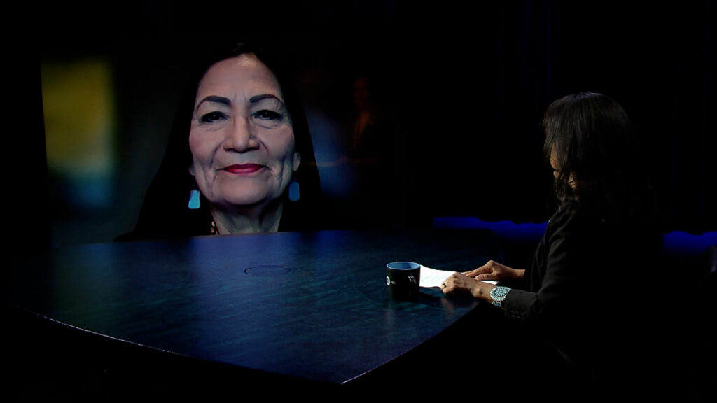 A woman sitting at a table in front of a television screen.