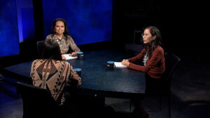 Three women sitting at a table in front of a blue screen.