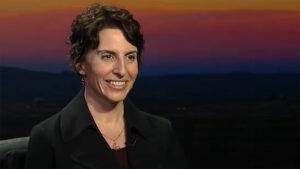 A woman in a black jacket smiling in front of a sunset.