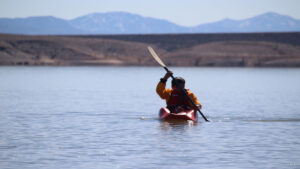A person is paddling a kayak in the water with mountains in the background.