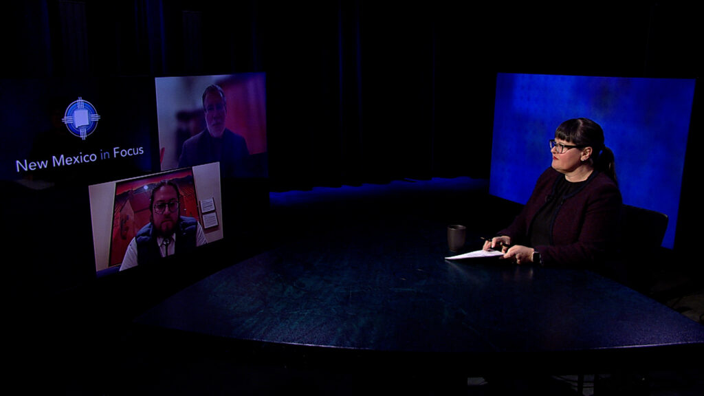 A woman sits at a table in front of two televisions.