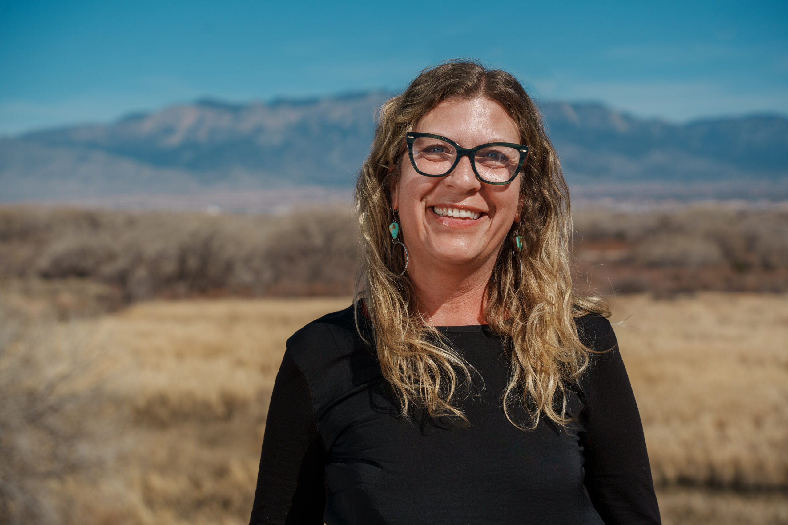 Smiling woman with glasses standing outdoors with a mountainous backdrop.