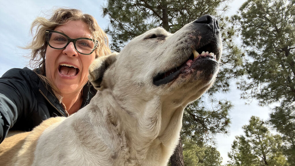 A woman with glasses is taking a selfie with her dog near the water.