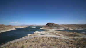 Clear skies over a large lake with a distinctive round hill and surrounding desert landscape.