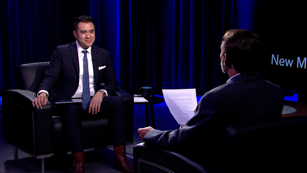 Two men in suits having a discussion on a television set, with one facing the camera and the other's back to the viewer.