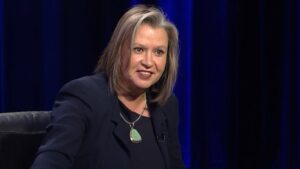 Woman with shoulder-length hair speaking during an interview, wearing a dark blazer and a necklace with a green pendant.