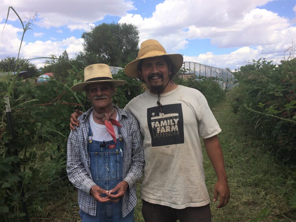 Two men in hats smiling and standing together in a garden with water and greenery in the background.