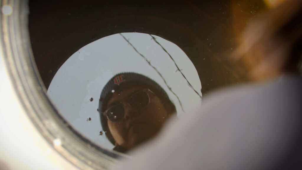 Reflection of a person wearing a beanie with a logo, seen in a round, sunlit surface, perhaps water, with a hint of blue sky and utility lines.
