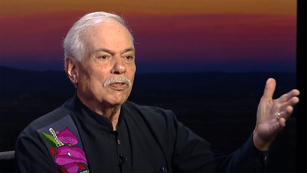 Elderly man with white hair and mustache wearing a black shirt, gesturing with his hand, seated in front of a sunset background.