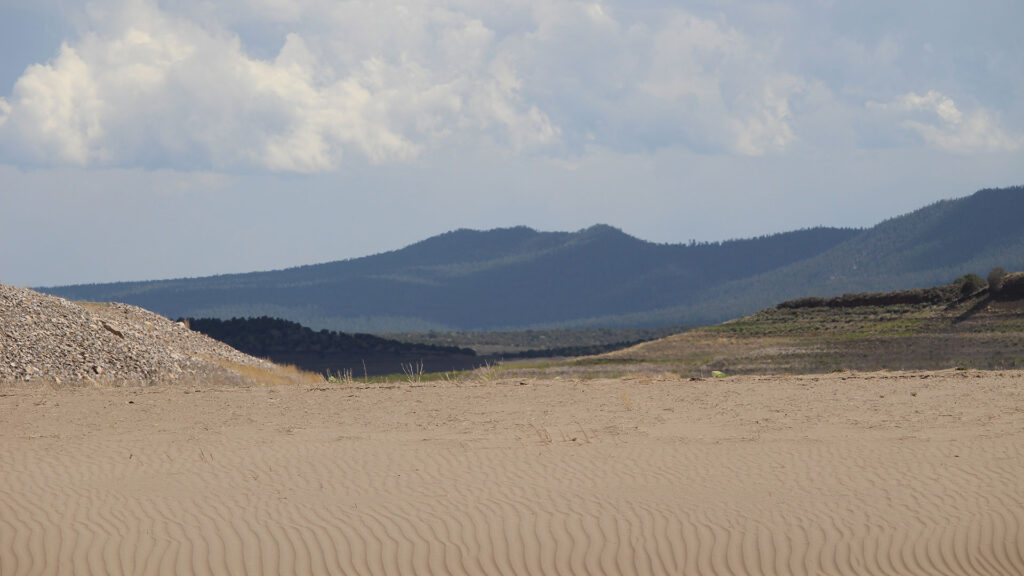 A desert landscape in the foreground with a mountain range under a cloudy sky and hints of water in the background.