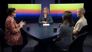 Four adults seated around a table in a tv studio setting, having a discussion, with a colorful abstract backdrop.