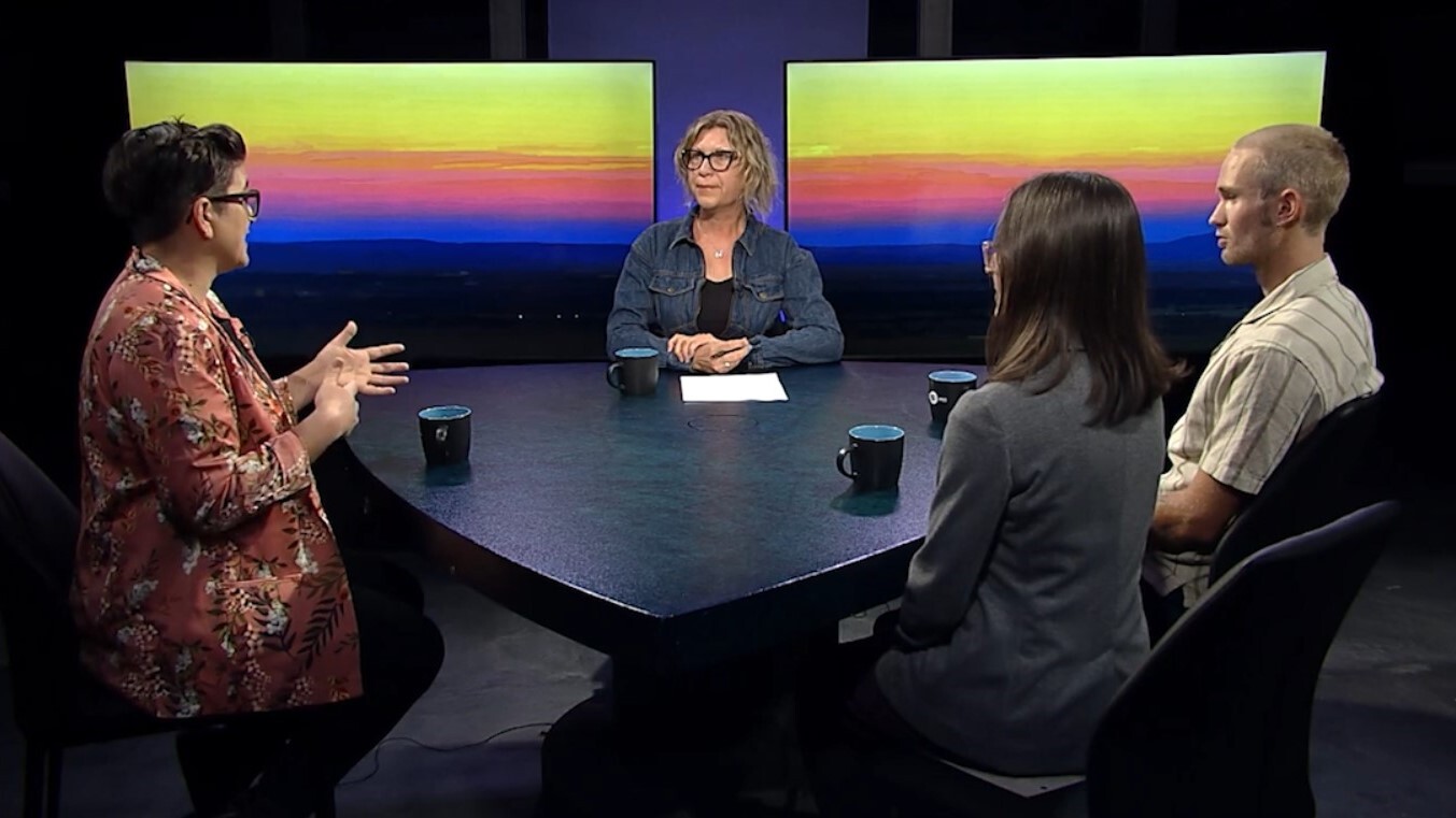 Four adults seated around a table in a tv studio setting, having a discussion, with a colorful abstract backdrop.