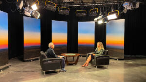 Two people seated in armchairs facing each other in a studio, with three large screens displaying a sunset landscape in the background.