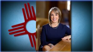 A smiling woman sitting at a table with the new mexico state flag in the background.