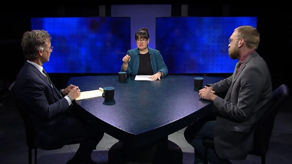 Two men and one woman engaged in a discussion at a round table on a talk show set.