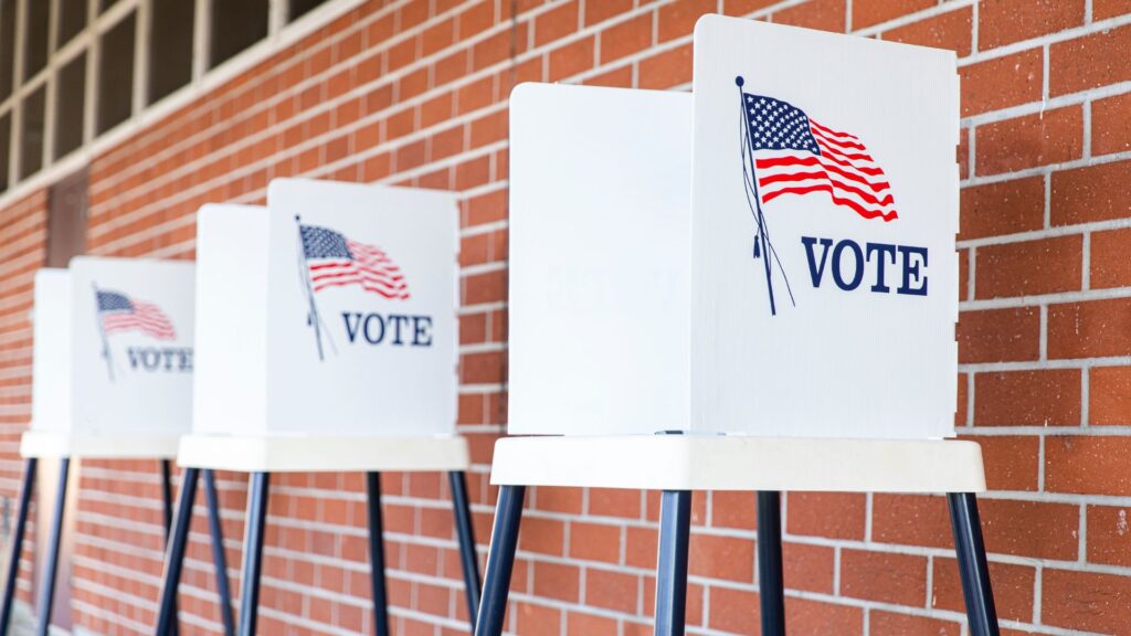 Four voting booths with american flags and the word "vote" on their screens, set against a brick wall background.