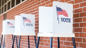 Four voting booths with american flags and the word "vote" on their screens, set against a brick wall background.