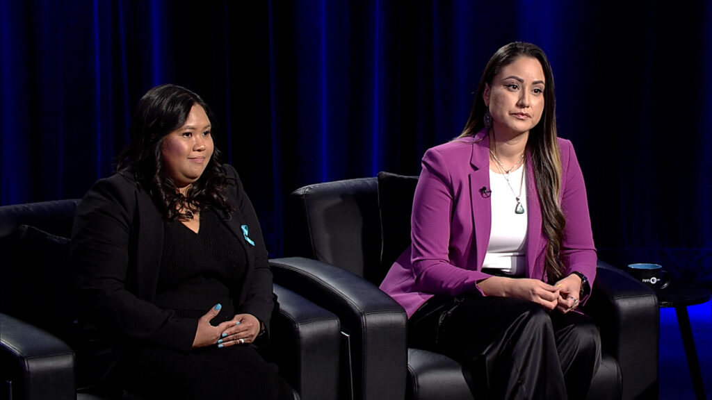 Two women sitting in armchairs on a stage set for a discussion, one wearing a teal dress and the other in a white blouse and purple blazer.