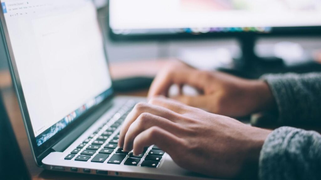 Close-up of hands typing on a laptop keyboard, with blurred dual monitors in the background.