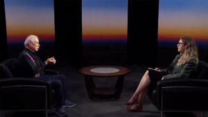Two people engaged in conversation on a tv set, with a sunset background. one person is an older man, the other a younger woman, both seated across a round table.