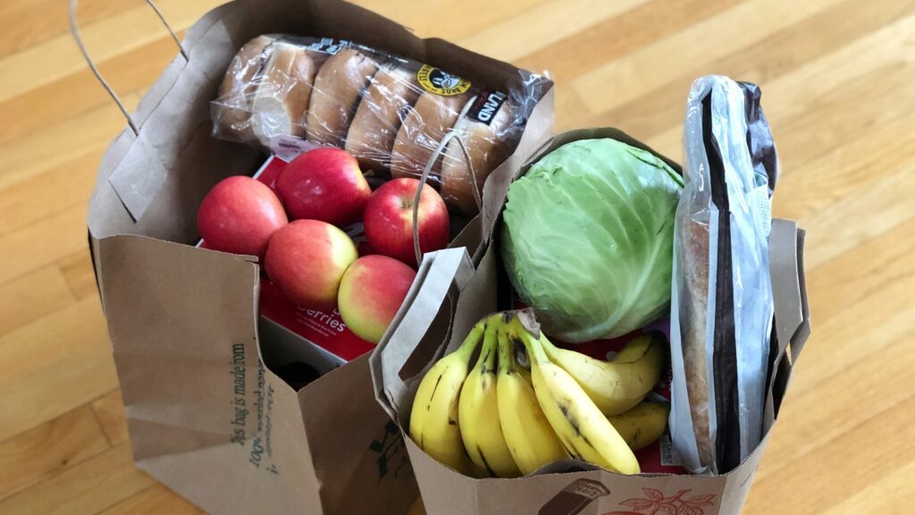 Two grocery bags on a wooden floor filled with fresh produce, including apples, bananas, and a cabbage, and packaged bread.