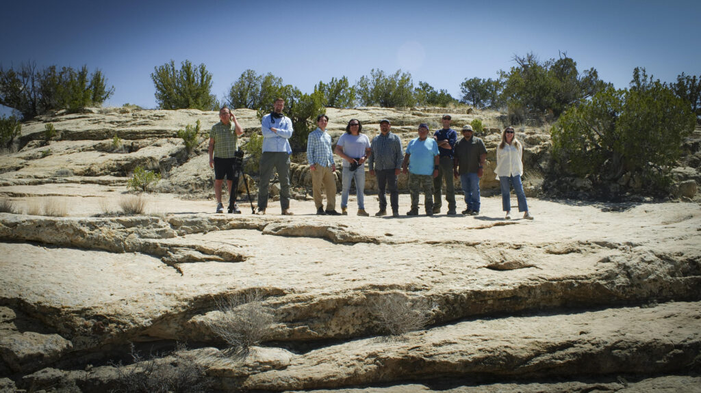 A group of nine adults standing on a rocky desert ledge near a body of water under a clear blue sky.