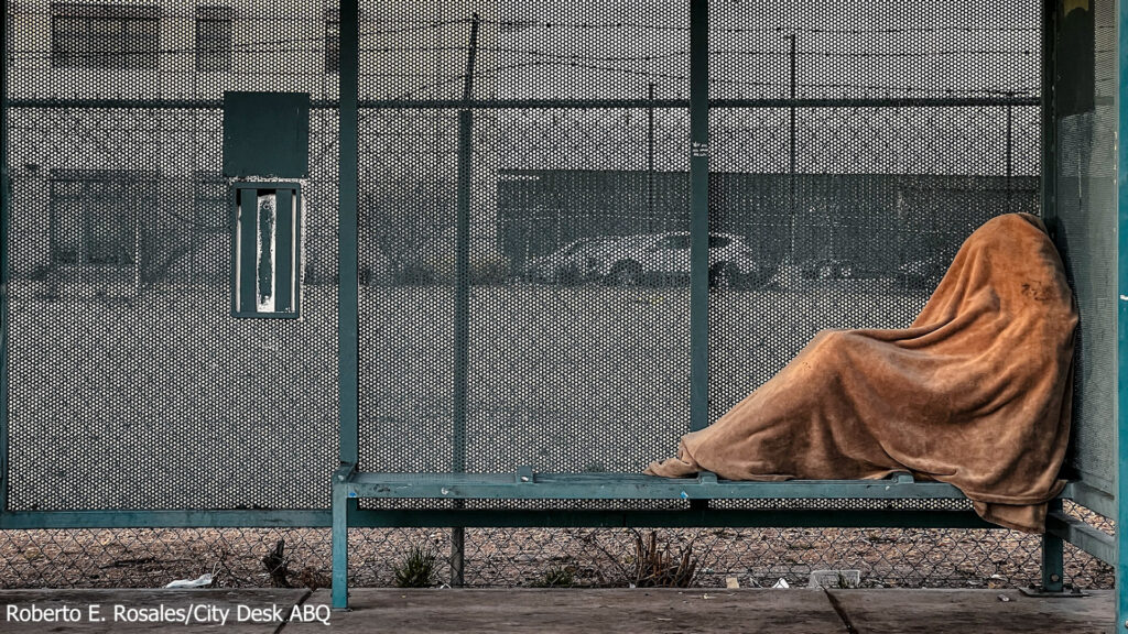 A person covered with a brown blanket sits on a green public bench, facing a chain-link fence with cars parked in the background.