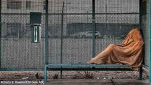 A person covered with a brown blanket sits on a green public bench, facing a chain-link fence with cars parked in the background.