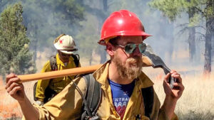 Two firefighters walk through a smoky forest, wearing protective gear and carrying tools. The firefighter in the foreground has a red helmet and sunglasses, while the other carries a hose ready to spray water at any hot spots.
