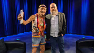 Two people posing under stage lights. The person on the left wears colorful clothing and holds a piece of paper. The person on the right has a beard and is dressed in a jacket and jeans.