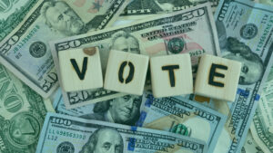 A variety of U.S. dollar bills serve as a backdrop for wooden blocks spelling out the word "VOTE.