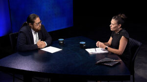 Two people sit at a table in a studio, engaged in conversation. Both have papers and coffee mugs in front of them. The background is dark with blue lighting on one side.