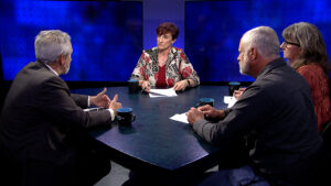 Four people engaged in a discussion around a table on a television set with a blue background.