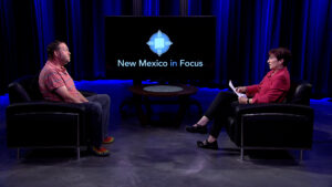 Two people, a man and a woman, sit in armchairs in a studio set for the tv show "new mexico in focus," facing each other across a small table.