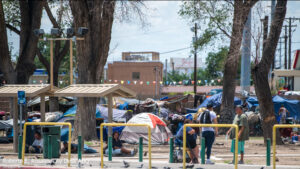 A busy urban park with people and tents scattered around, some sitting and walking, amidst green trees and playground equipment.