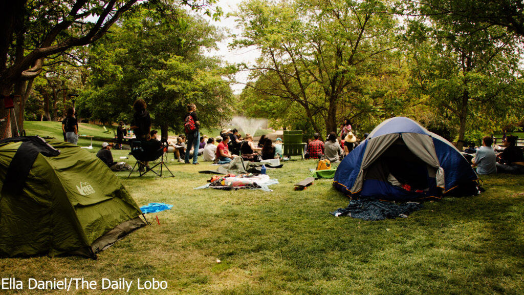 Group of people socializing in a park with tents, blankets, and a barbecue grill, surrounded by trees.