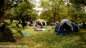 Group of people socializing in a park with tents, blankets, and a barbecue grill, surrounded by trees.