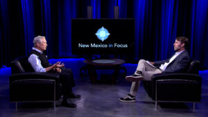 Two men having a discussion on a tv show set with the title "new mexico in focus" displayed on a screen behind them.