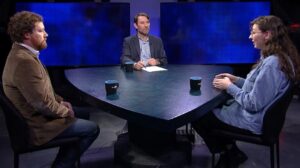 Three people engaged in a discussion around a triangular table with two mugs. The middle person has a piece of paper in front of him. The background consists of three dark monitors.